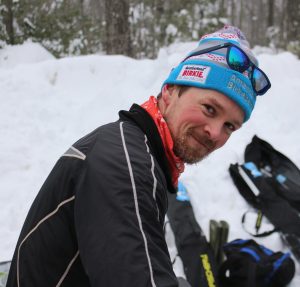 Assistant Coach Sam Holmes in light blue beanie with sunglasses perched on top in front of ski bags and a snowbank