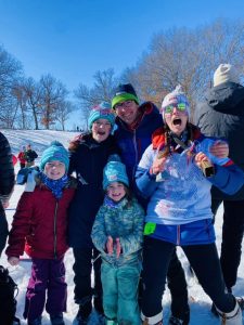 A family in snow pants and jackets looks at the camera in front of a snowy hill with bare trees